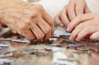 Zdjęcie przedstawia dłonie seniora oraz młodej osoby układające puzzle