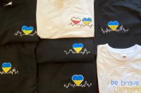 na zdjęciu są czarne i białe koszulki z logotypem w kształcie serca. W środku logotypu kolory Ukrainy i Polski