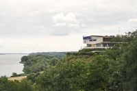 Na zdjęciu Dom turysty PTTK. Domek letniskowy na wybrzerzu. Na pierwszym planie rołśinność nadmorska, oraz fragment kamiennego nadbrzeża. W tle morze oraz panorama nieba
