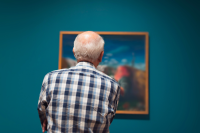 Senior w muzeum patrzy na obraz. Ubrany jest w koszulę w kratę. W tle obraz wiszący na turkosowej ścianie.  ścianie 