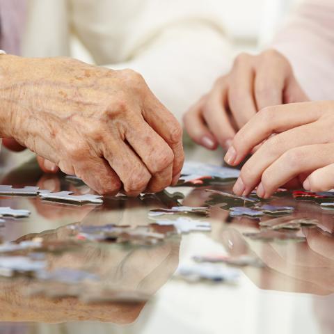 Zdjęcie przedstawia dłonie seniora oraz młodej osoby układające puzzle