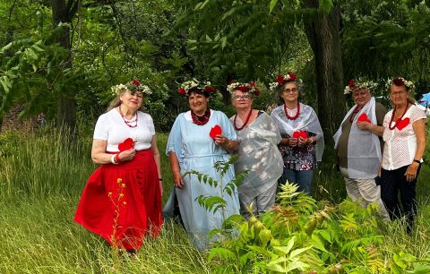 Szóstka Pań z grupy Aktywne Wrocławianki pozuje do zdjęcia. Panie stoją przed lasem, wśród zieleni i drzew. Mają na głowach kwietne wianki, na szyji czerwone korale. oraz przyczepione ręcznie robione serca przypięte do bluzek. 