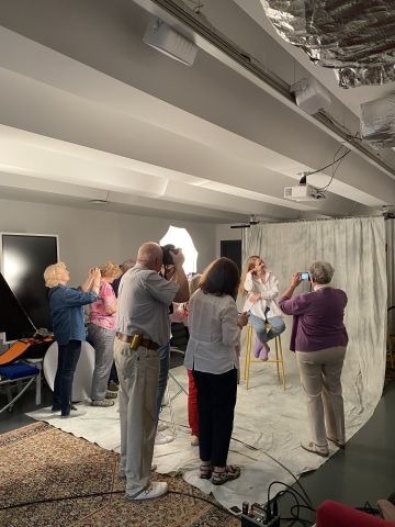Na zdjęciu w zaaranżowanym studio grupa kilku seniorów robi zdjęcie pozującej modelce, która siedzi wysokim stołku