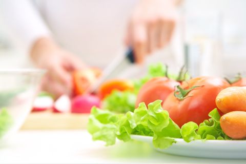 na zdjęciu na pierwszym planie mamy talerz a nim warzywa: sałata, pomidor, marchewka. W tle rozmyte ręce osoby, która kroi warzywa