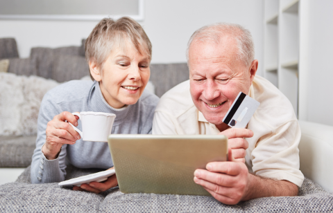 Na zdjęciu w pokoju uśmiechnięta para seniorów przy laptopie z kartą płatniczą.  
