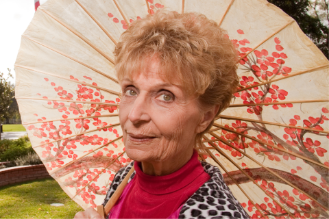 Zdjęcie przedstawia kobietę pod parasolem w słoneczny dzień. W tle widoczny fragment parku z zielenią