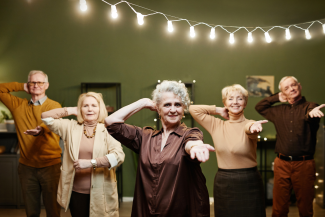 % osób w wieku seniorlanym tańczy w pokoju. U góry zdjęcia widoczne są roświetlone w szeregu żarówki.