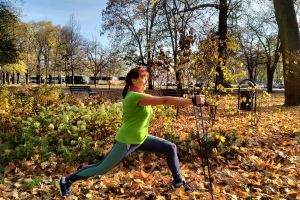 Na zdjęciu widzimy jesienny park i seniorkę, która stoi bokiem w dużym rozkroku i wykonuje ćwiczenie z kilkami