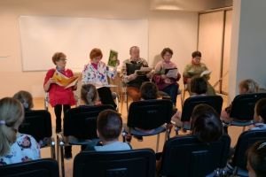 Seniorzy siedzący na krzesłach w bibliotece czytający bajki dzieciom w ramach projektu Poczytajki Babci i Dziadka.