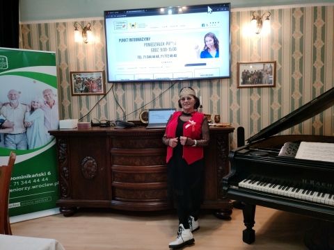 Na zdjęciu kobieta w kolorowym ubraniu w sali wykładowej przy fortepianie.