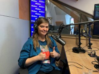 Na zdjęciu w studiu radiowym dziewczyna z uniesionymi kciukami do góry. 