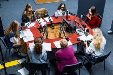 Na zdjęciu przy stole osoby w różnym wieku przy mikrofonach.