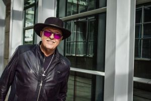 Zdjęcie przedstawia muzyka Andrzeja Cierniewskiego, który ubrany jest w czarną skórzaną kurtkę, czarny kapelusz oraz okulary z fioletowymi szkłami. Artysta pozuje na tle szklanych okien biurowca