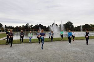 Grupa seniorów podczas ćwiczeń z kijkami nordic walking w parku przy fontannie na terenie przyległym do hali stulecia. 