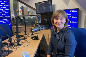 Na zdjęciu kobieta siedząca przy stole i przy mikrofonie w małym pomieszczeniu.