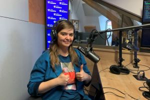Na zdjęciu w studiu radiowym dziewczyna z uniesionymi kciukami do góry. 